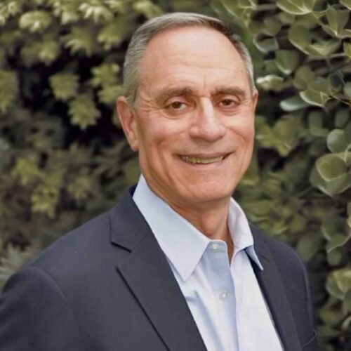 Sandy Alexander, a middle-aged man with short gray hair, wears a dark blazer and light blue shirt, smiling warmly in front of lush green foliage.
