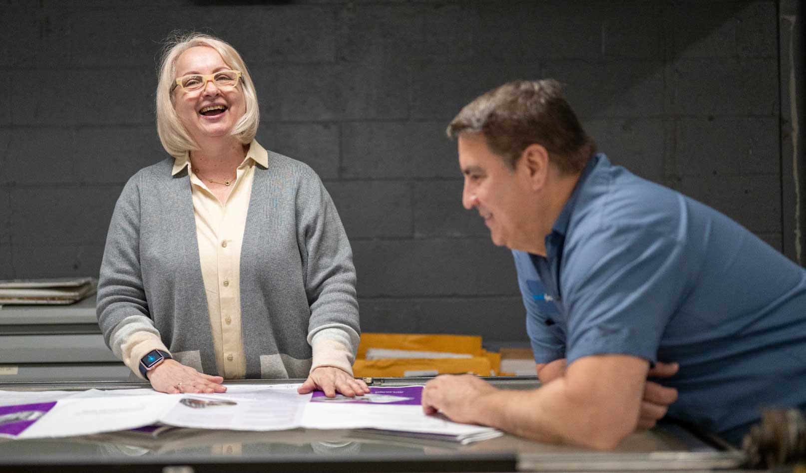 Two people stand at a table covered with papers, smiling and talking. The woman on the left, possibly Sandy Alexander, is laughing and wearing glasses; the man on the right listens in a blue shirt. A plain wall forms the background.