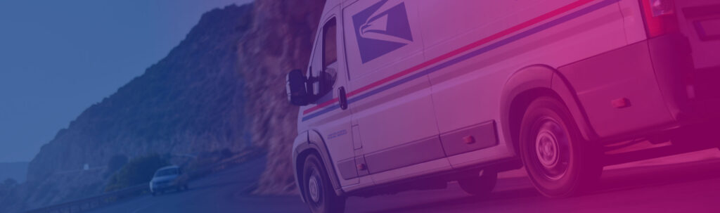 A Sandy Alexander postal delivery van with red and blue stripes drives on a winding road next to a rocky hillside, with another vehicle following behind.