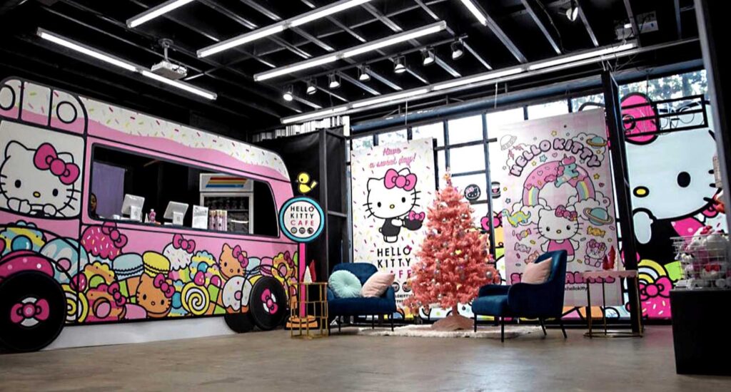 A colorful Hello Kitty-themed café designed by Sandy Alexander, featuring a pink Christmas tree, blue chairs, and cartoon wall art with Hello Kitty and sweets for a playful and vibrant atmosphere.