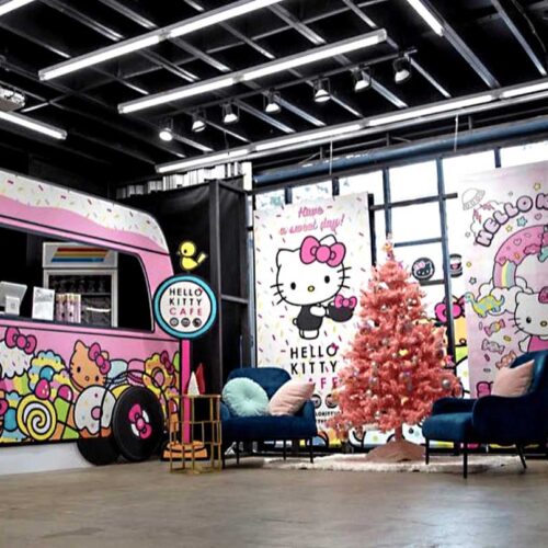 A colorful Hello Kitty-themed café designed by Sandy Alexander, featuring a pink Christmas tree, blue chairs, and cartoon wall art with Hello Kitty and sweets for a playful and vibrant atmosphere.