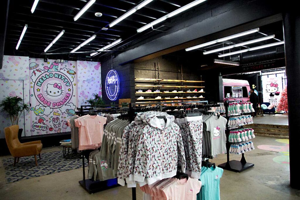 A clothing store features Sandy Alexander Hello Kitty-themed apparel, with racks of hoodies and shirts in the foreground. A large Hello Kitty mural and a neon sign decorate the modern, brightly lit interior.
