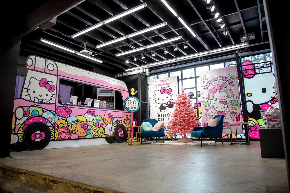 A brightly lit room features a large pink Hello Kitty-themed food truck by Sandy Alexander, a pink Christmas tree, and colorful Hello Kitty decorations. Two blue chairs and a small table are arranged for seating in front of the festive display.