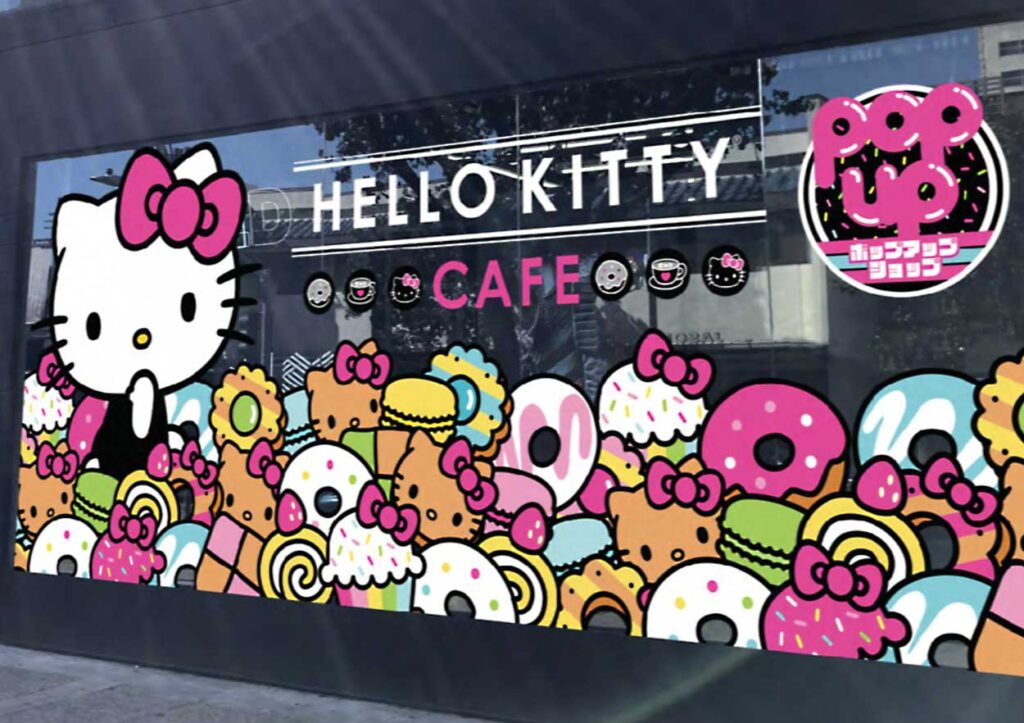 Storefront window decorated by Sandy Alexander with Hello Kitty and colorful cartoon desserts, featuring the text HELLO KITTY CAFE and Pop Up in pink. The whimsical design includes donuts, cupcakes, macarons, and confetti.