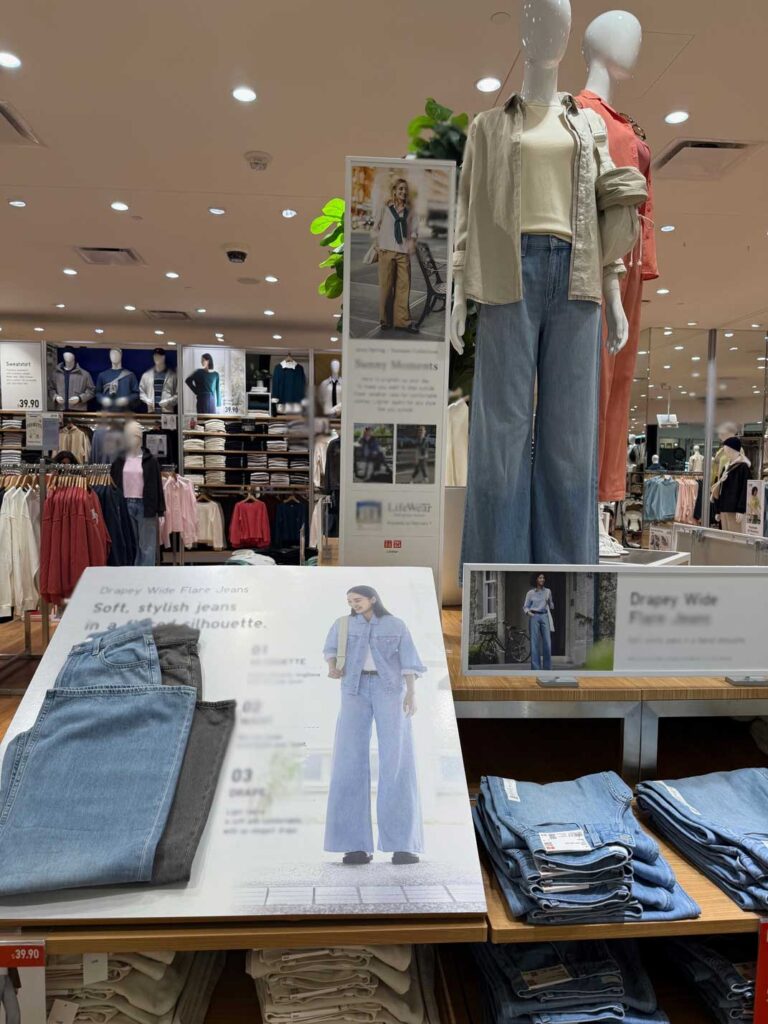 A clothing store display features mannequins in wide-leg jeans and shirts, with folded jeans on tables below and posters of Sandy Alexander modeling similar outfits. The store is brightly lit and neatly organized.