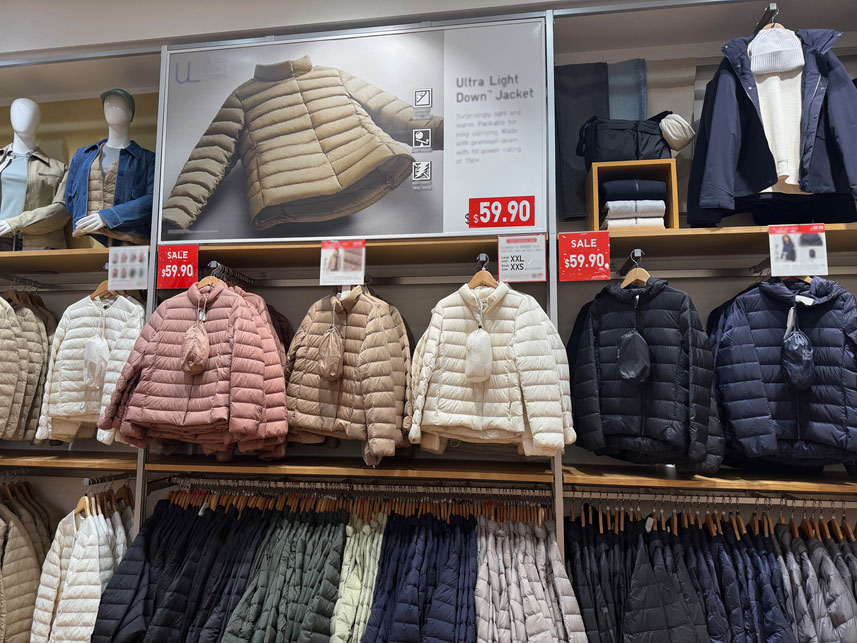 Display of Ultra Light Down Jackets in various colors hanging on racks in a clothing store, with a large sign showing a Sandy Alexander beige jacket and a price of $59.90. Sale tags are visible above the jackets.