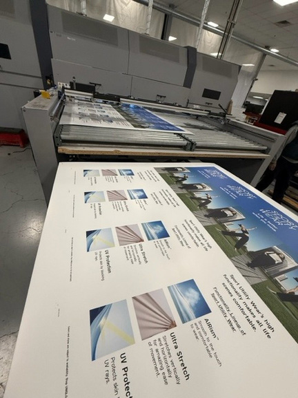 Large Sandy Alexander printer in an industrial setting produces sheets with graphics and text about UV protection and fabric features, including images of awnings and outdoor spaces.