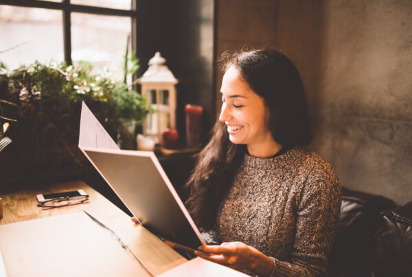 A woman with long dark hair sits at a wooden table by a window, smiling while looking at a menu in a cozy, warmly lit restaurant with potted plants and candles in the background.