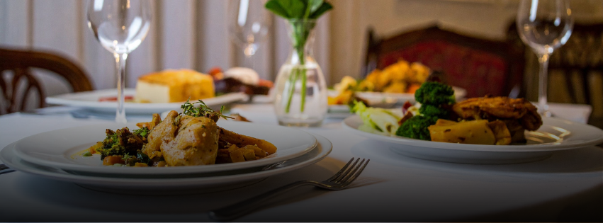A close-up of a dining table set with plates of gourmet food, wine glasses, and a small vase with green flowers in the background, creating an elegant restaurant atmosphere.