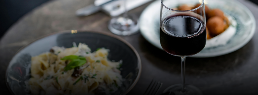 A glass of red wine sits on a table next to a plate of pasta and another plate of round appetizers, with cutlery and a napkin in the background.