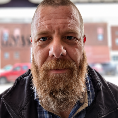 Bearded man with short hair wearing a plaid shirt and dark jacket, standing outdoors with a blurred background of buildings and parked cars.