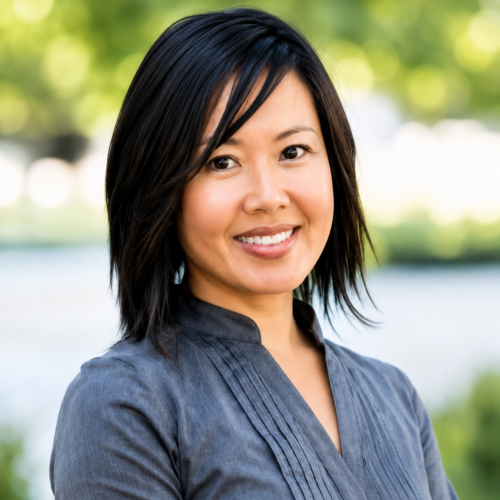 A woman with straight, shoulder-length black hair smiles outdoors. She wears a gray blouse and stands in front of a blurred, green background with sunlight filtering through trees.