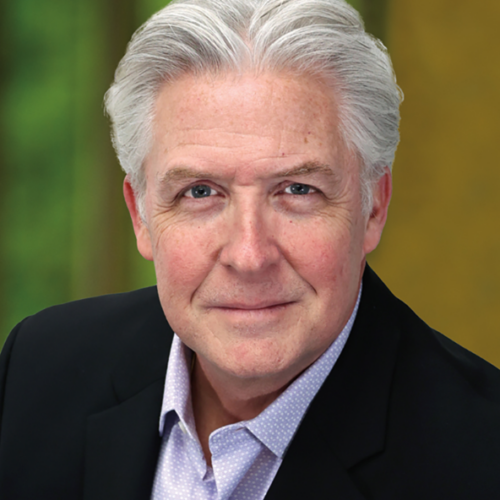 Smiling older man with white hair wearing a black blazer and light purple shirt, posing in front of a blurred green and yellow background.