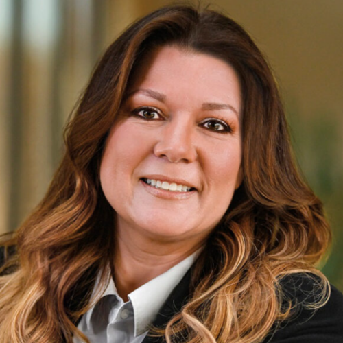 Smiling woman with long wavy brown hair, wearing a black blazer and light-colored collared shirt, posed in an indoor setting with a blurred background.